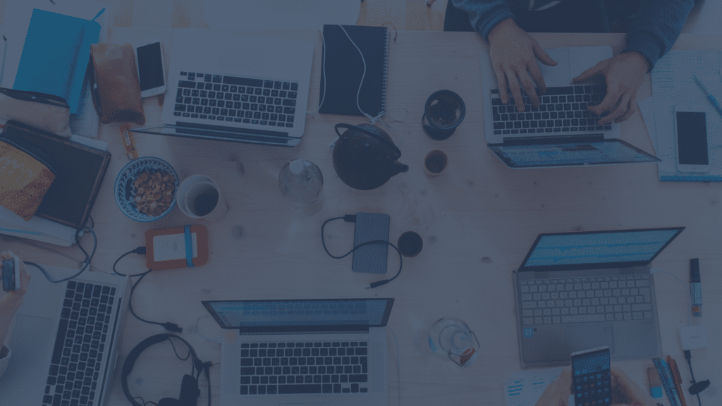 An overhead, flat-lay view of a wooden table covered in laptops, smartphones, tech accessories, and coffee cups, with a dark blue color overlay. Hands likely belong to B2B SaaS PR professionals.